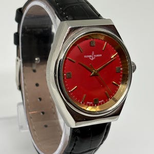 May include: An Ulysse Nardin wristwatch featuring a vibrant red dial with gold-tone hour markers and hands. The watch has a silver-tone metal case and a black leather strap with a textured, embossed pattern. The text Ulysse Nardin and Swiss Made are visible on the dial.