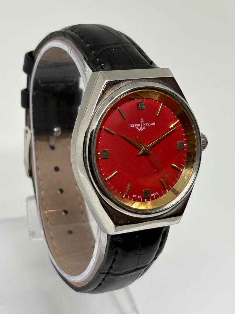 May include: An Ulysse Nardin wristwatch featuring a vibrant red dial with gold-tone hour markers and hands. The watch has a silver-tone metal case and a black leather strap with a textured, embossed pattern. The text Ulysse Nardin and Swiss Made are visible on the dial.