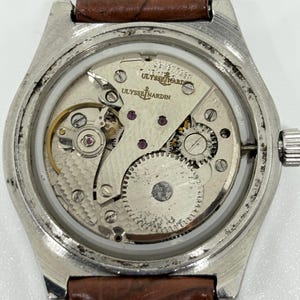 May include: A silver-toned wristwatch with an exposed mechanical movement visible through the case back. The movement features the Ulysse Nardin logo and anchor emblem. The watch is attached to a brown leather strap with visible white stitching.