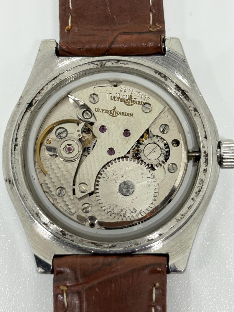 May include: A silver-toned wristwatch with an exposed mechanical movement visible through the case back. The movement features the Ulysse Nardin logo and anchor emblem. The watch is attached to a brown leather strap with visible white stitching.