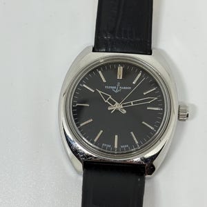 May include: A silver-tone Ulysse Nardin wristwatch with a round black dial, silver hour markers, and silver hands. The watch features a black textured leather strap and a polished metal case. The dial displays the Ulysse Nardin logo and Swiss Made.
