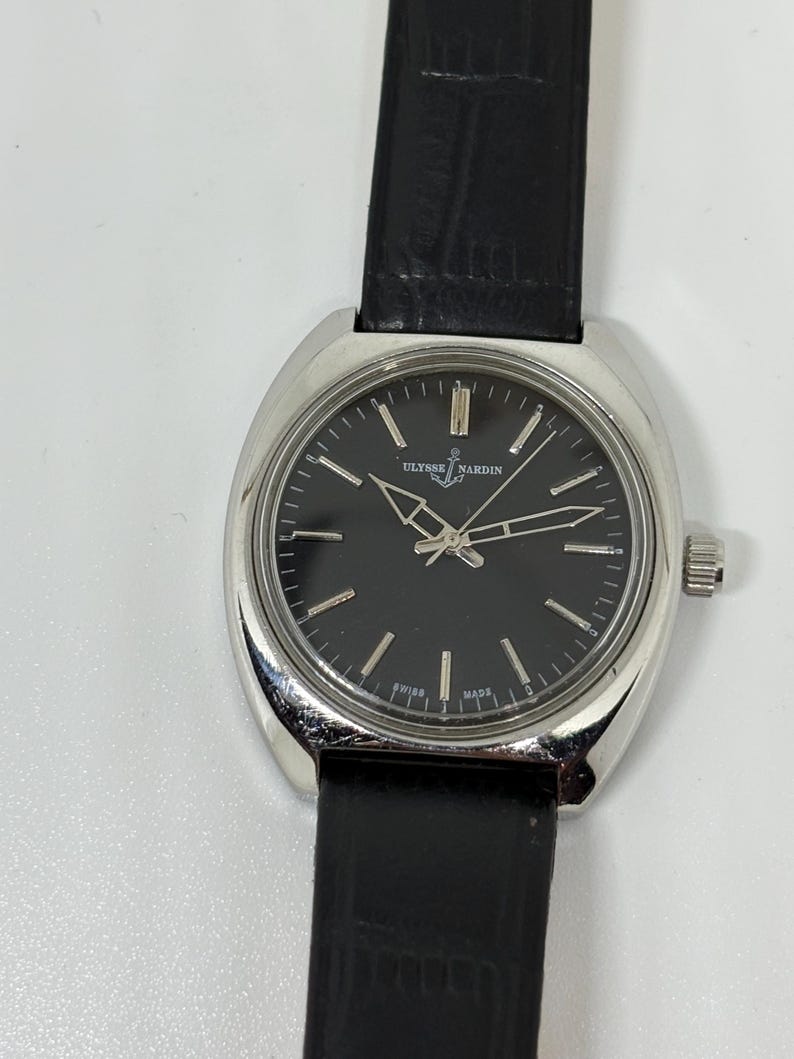 May include: A silver-tone Ulysse Nardin wristwatch with a round black dial, silver hour markers, and silver hands. The watch features a black textured leather strap and a polished metal case. The dial displays the Ulysse Nardin logo and Swiss Made.
