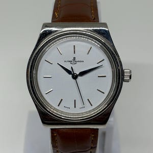 May include: A silver-toned wristwatch with a white dial, featuring black hour markers and hands. The watch face displays the text Ulysse Nardin with an anchor logo and the words Swiss Made. It is attached to a brown leather strap with a textured finish.