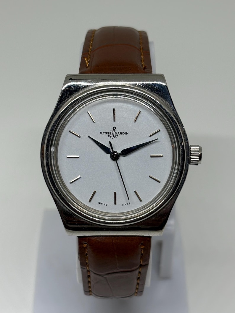 May include: A silver-toned wristwatch with a white dial, featuring black hour markers and hands. The watch face displays the text Ulysse Nardin with an anchor logo and the words Swiss Made. It is attached to a brown leather strap with a textured finish.