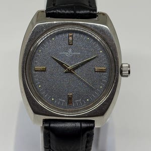 Może przedstawiać: Zegarek na rękę Ulysse Nardin z teksturowaną ciemnoszarą tarczą, złotymi znacznikami godzin i wskaz&oacute;wkami. Zegarek posiada srebrną kopertę ze stali nierdzewnej oraz czarny sk&oacute;rzany pasek z tłoczonym wzorem krokodyla i srebrną klamrą.