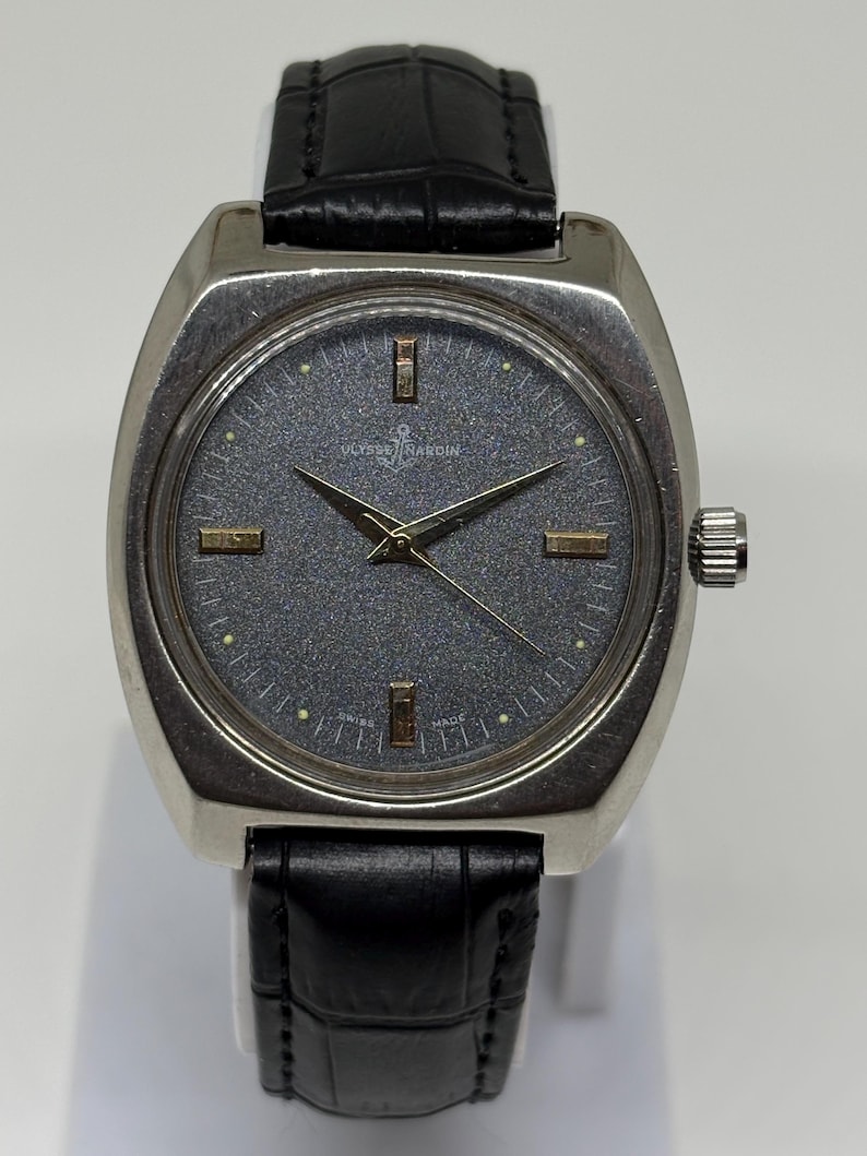 Może przedstawiać: Zegarek na rękę Ulysse Nardin z teksturowaną ciemnoszarą tarczą, złotymi znacznikami godzin i wskaz&oacute;wkami. Zegarek posiada srebrną kopertę ze stali nierdzewnej oraz czarny sk&oacute;rzany pasek z tłoczonym wzorem krokodyla i srebrną klamrą.