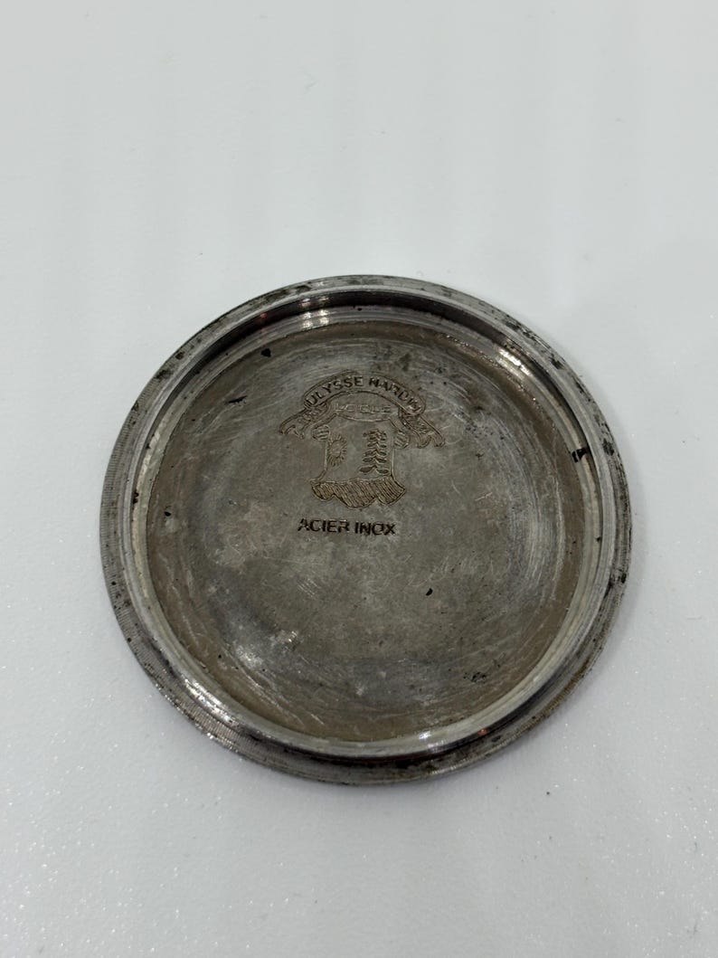 May include: A circular silver-toned metal watch case back featuring an engraved crest with the text Ulysse Nardin and the words ACIER INOX stamped below the emblem. The metallic surface shows signs of wear and age with a polished rim.