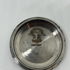 May include: A circular silver-toned metal watch case back with a polished finish. The interior features an engraved crest logo with the text ULYSSE NARDIN and the words ACIER INOX printed below the emblem in a clear, dark font.