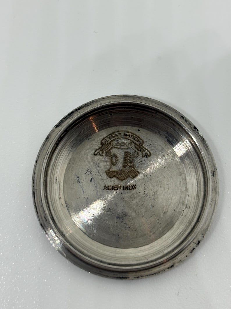 May include: A circular silver-toned metal watch case back with a polished finish. The interior features an engraved crest logo with the text ULYSSE NARDIN and the words ACIER INOX printed below the emblem in a clear, dark font.