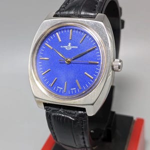 May include: A vintage Ulysse Nardin watch with a square silver case and a vibrant blue dial. The watch features gold-tone hands and markers, a black leather strap, and the brand's logo. The watch is displayed on a black and red stand.