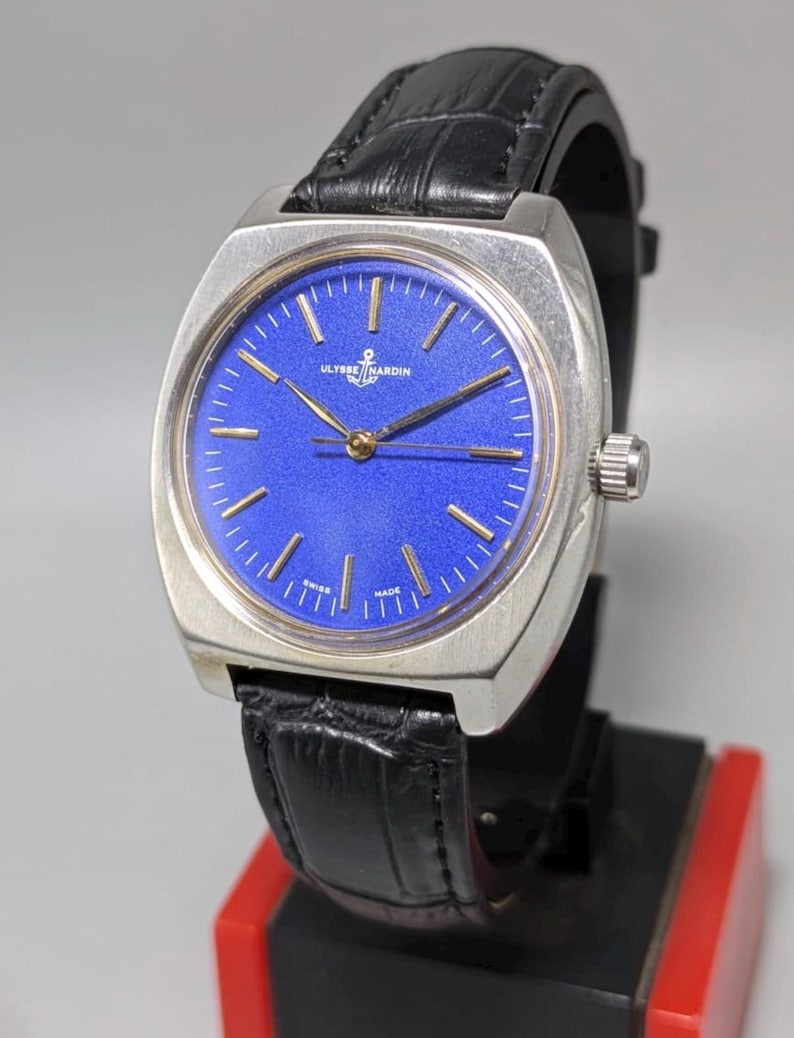 May include: A vintage Ulysse Nardin watch with a square silver case and a vibrant blue dial. The watch features gold-tone hands and markers, a black leather strap, and the brand's logo. The watch is displayed on a black and red stand.