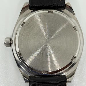 May include: A silver-toned wristwatch with a circular case back featuring a radial brushed metal finish. The watch includes a black leather strap with visible white stitching and a small, ridged winding crown on the left side of the case.