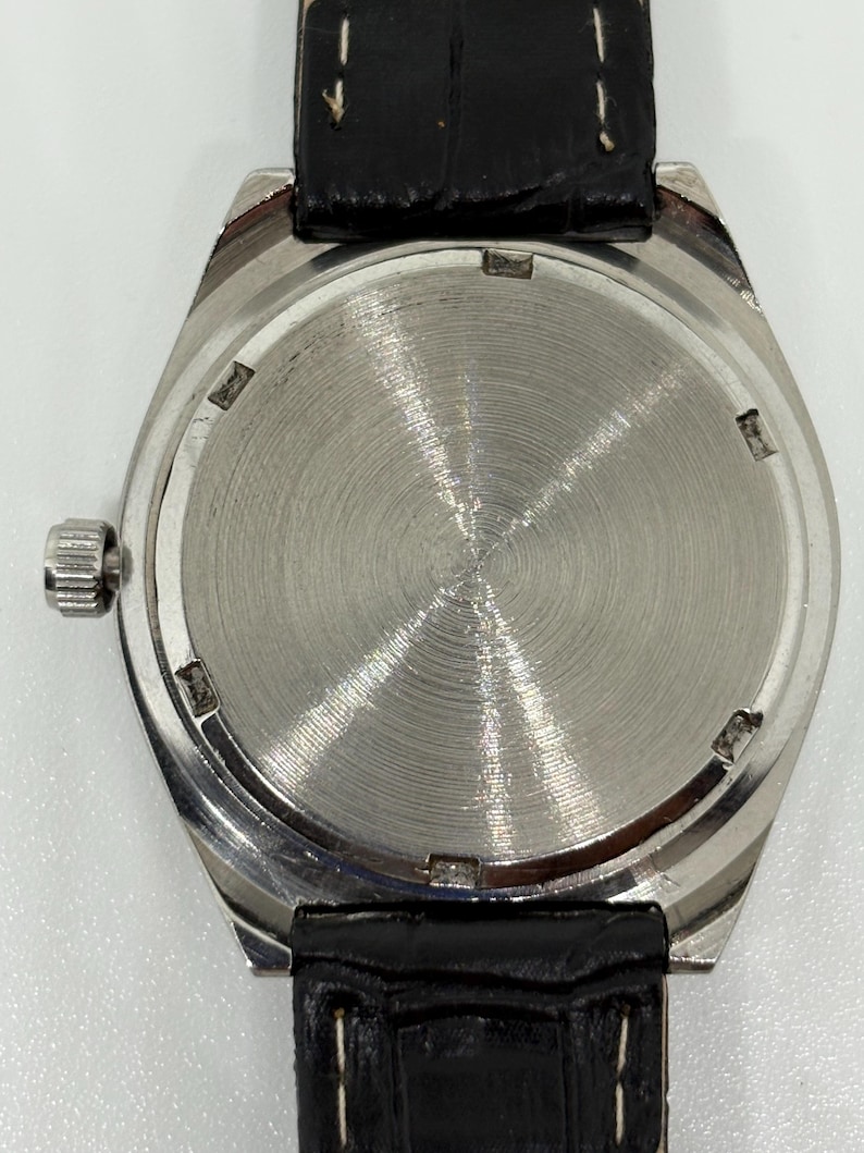 May include: A silver-toned wristwatch with a circular case back featuring a radial brushed metal finish. The watch includes a black leather strap with visible white stitching and a small, ridged winding crown on the left side of the case.