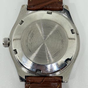 May include: A silver-toned watch case with a circular, brushed metal back featuring concentric grooves. The watch is attached to a brown, textured leather strap with white stitching. A small, ridged metal crown is visible on the left side.