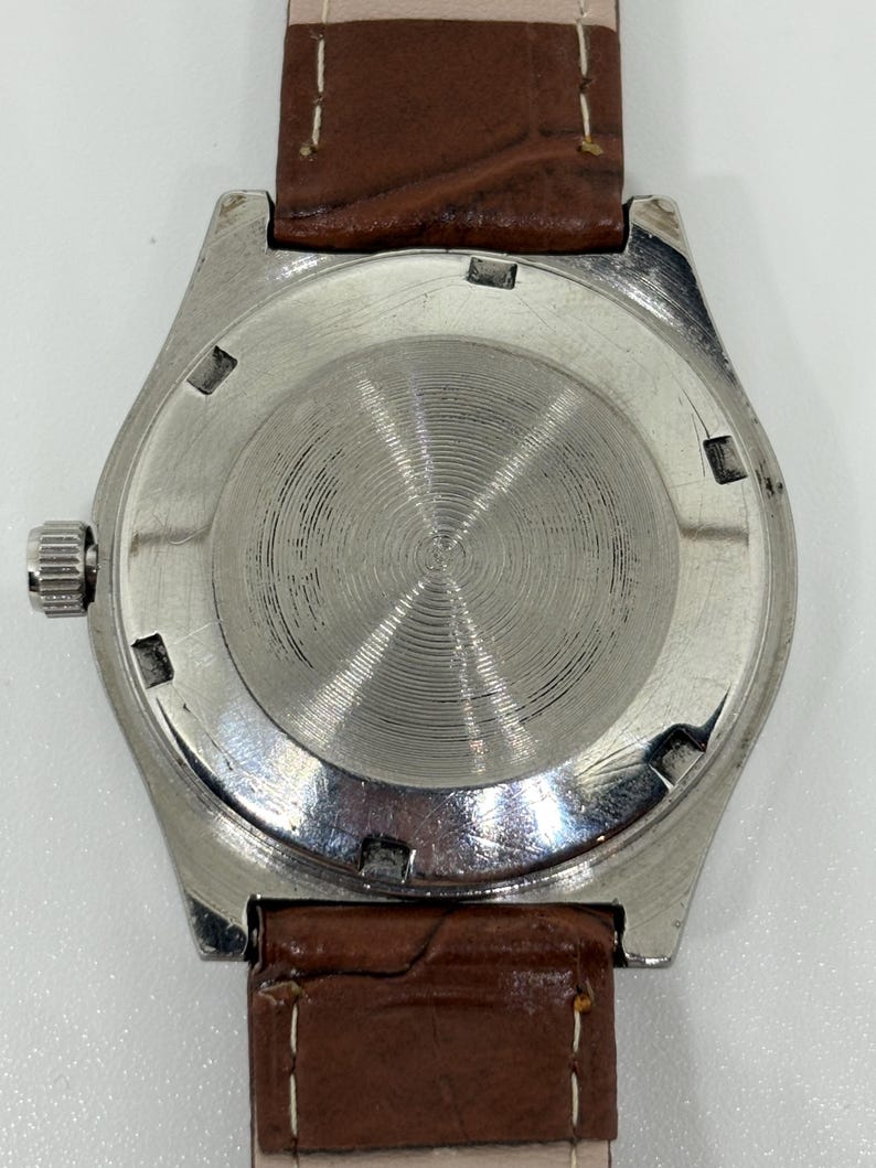 May include: A silver-toned watch case with a circular, brushed metal back featuring concentric grooves. The watch is attached to a brown, textured leather strap with white stitching. A small, ridged metal crown is visible on the left side.