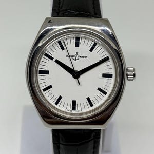 May include: A stainless steel Ulysse Nardin wristwatch with a white dial, black hour markers, and black hands. The watch features a black leather strap with a textured finish and a silver-toned crown on the right side of the case.