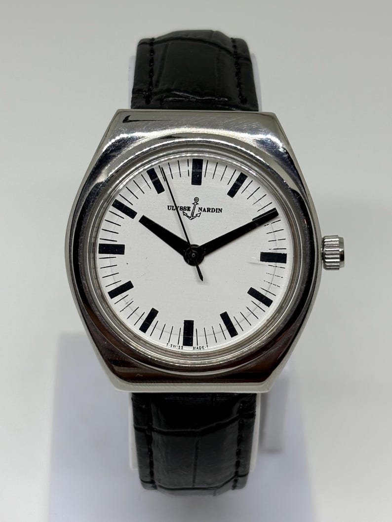 May include: A stainless steel Ulysse Nardin wristwatch with a white dial, black hour markers, and black hands. The watch features a black leather strap with a textured finish and a silver-toned crown on the right side of the case.