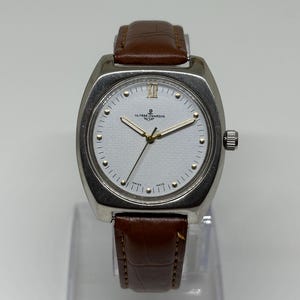 K&ouml;nnte beinhalten: Eine silberfarbene Armbanduhr mit wei&szlig;em Zifferblatt, goldfarbenen Zeigern und einem braunen Lederarmband mit strukturierter Oberfl&auml;che. Das Zifferblatt zeigt das Ulysse Nardin-Logo, ein Ankersymbol und den Text Swiss Made. Oben befindet sich die r&ouml;mische XII.