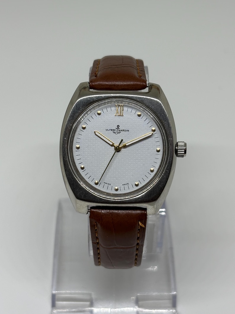K&ouml;nnte beinhalten: Eine silberfarbene Armbanduhr mit wei&szlig;em Zifferblatt, goldfarbenen Zeigern und einem braunen Lederarmband mit strukturierter Oberfl&auml;che. Das Zifferblatt zeigt das Ulysse Nardin-Logo, ein Ankersymbol und den Text Swiss Made. Oben befindet sich die r&ouml;mische XII.