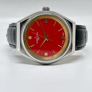 May include: A wristwatch with a vibrant red dial, gold-tone hands, and gold-tone hour markers. The watch features a silver-tone metal case and a black leather strap with a textured finish. The text Ulysse Nardin and Swiss Made are visible.