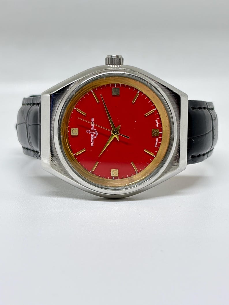 May include: A wristwatch with a vibrant red dial, gold-tone hands, and gold-tone hour markers. The watch features a silver-tone metal case and a black leather strap with a textured finish. The text Ulysse Nardin and Swiss Made are visible.