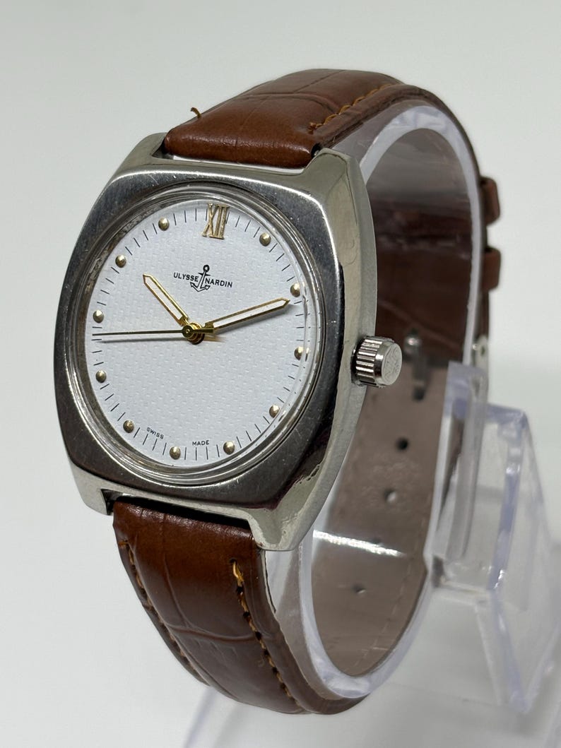 K&ouml;nnte beinhalten: Eine Armbanduhr mit einem silberfarbenen kissenf&ouml;rmigen Geh&auml;use und einem wei&szlig;en, strukturierten Zifferblatt. Das Zifferblatt zeigt goldfarbene Stundenmarkierungen, eine r&ouml;mische Ziffer auf der Zw&ouml;lf und den Schriftzug Ulysse Nardin mit Anker-Logo.