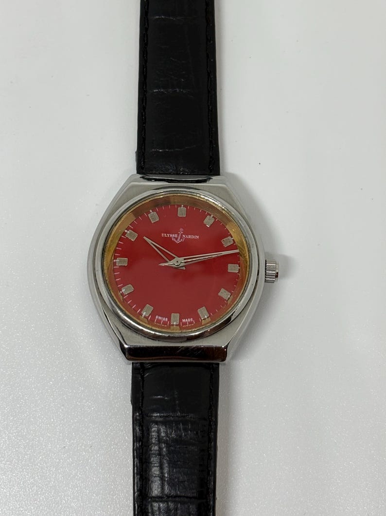 May include: A wristwatch with a circular silver-tone case, a vibrant red dial, and silver-tone hour markers. The dial features the text Ulysse Nardin and Swiss Made. It is attached to a black leather strap with a textured, crocodile-embossed finish.