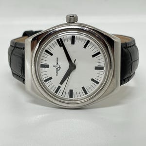 May include: A silver-toned wristwatch with a round white dial, black hour markers, and black hands. The watch features the Ulysse Nardin logo and anchor symbol on the face, with a black textured leather strap and a silver crown at the top position.