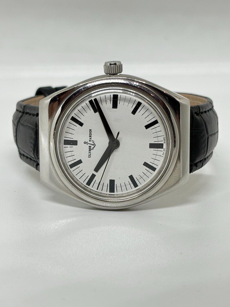 May include: A silver-toned wristwatch with a round white dial, black hour markers, and black hands. The watch features the Ulysse Nardin logo and anchor symbol on the face, with a black textured leather strap and a silver crown at the top position.