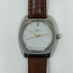 K&ouml;nnte beinhalten: Eine silberfarbene Armbanduhr mit wei&szlig;em Zifferblatt, goldfarbenen Zeigern und goldfarbenen Stundenmarkierungen. Die Uhr tr&auml;gt den Schriftzug Ulysse Nardin mit einem Anker-Logo und Swiss Made am unteren Rand. Sie hat ein braunes Lederarmband.