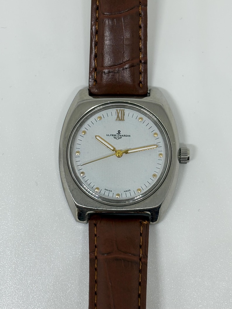 K&ouml;nnte beinhalten: Eine silberfarbene Armbanduhr mit wei&szlig;em Zifferblatt, goldfarbenen Zeigern und goldfarbenen Stundenmarkierungen. Die Uhr tr&auml;gt den Schriftzug Ulysse Nardin mit einem Anker-Logo und Swiss Made am unteren Rand. Sie hat ein braunes Lederarmband.