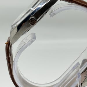 K&ouml;nnte beinhalten: Eine Seitenansicht einer Armbanduhr mit einem polierten silberfarbenen Metallgeh&auml;use und einem braunen, strukturierten Lederarmband. Die Uhr verf&uuml;gt &uuml;ber eine kleine, runde Krone an der Seite mit einer sichtbaren Logogravur und ein gew&ouml;lbtes Glas.