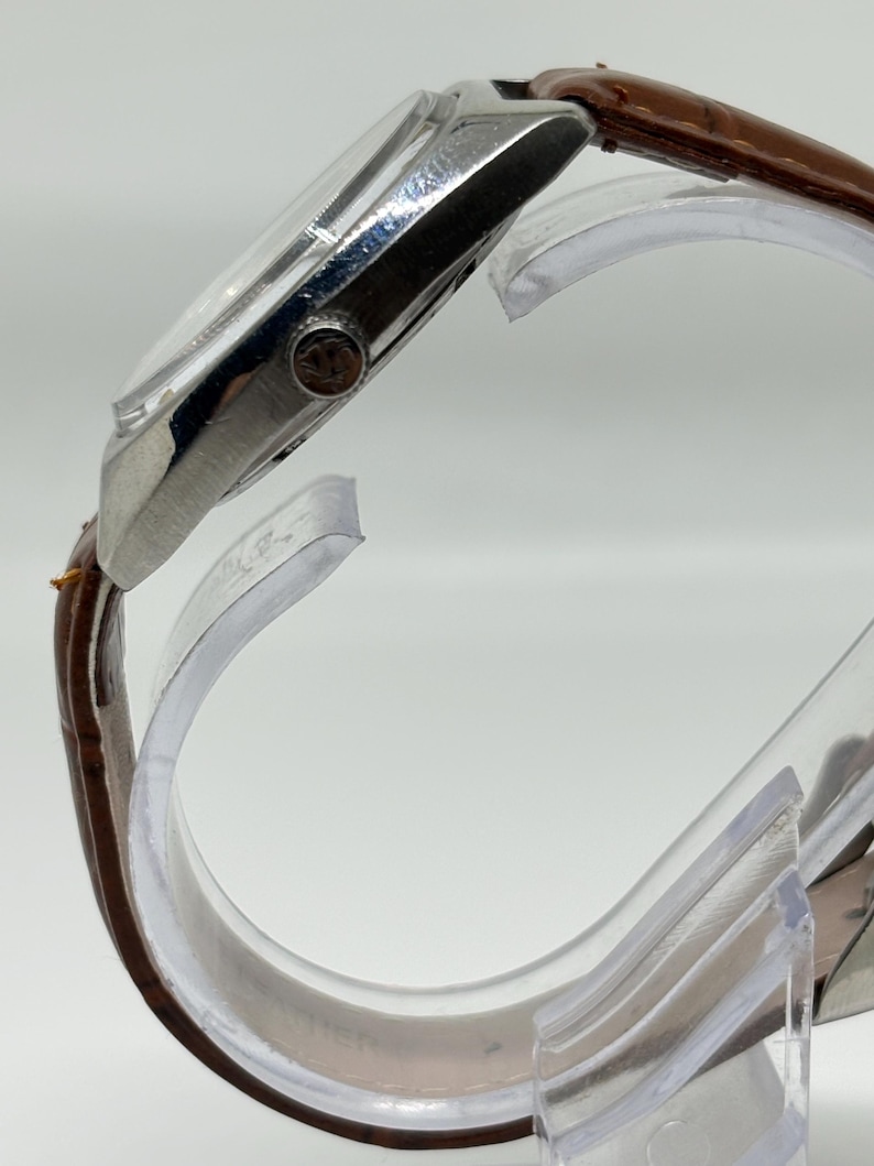 K&ouml;nnte beinhalten: Eine Seitenansicht einer Armbanduhr mit einem polierten silberfarbenen Metallgeh&auml;use und einem braunen, strukturierten Lederarmband. Die Uhr verf&uuml;gt &uuml;ber eine kleine, runde Krone an der Seite mit einer sichtbaren Logogravur und ein gew&ouml;lbtes Glas.