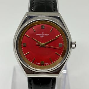 May include: A Ulysse Nardin wristwatch featuring a vibrant red dial with gold-tone hour markers and hands. The watch has a silver-tone metal case and a black leather strap with a textured finish. The text Ulysse Nardin and Swiss Made are visible.