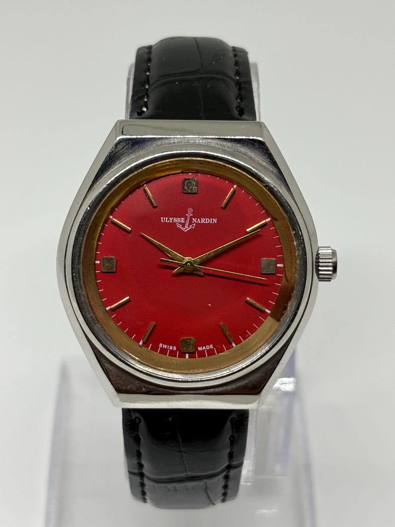 May include: A Ulysse Nardin wristwatch featuring a vibrant red dial with gold-tone hour markers and hands. The watch has a silver-tone metal case and a black leather strap with a textured finish. The text Ulysse Nardin and Swiss Made are visible.