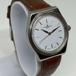 May include: A silver-tone wristwatch with a white dial featuring the Ulysse Nardin logo and anchor symbol. The watch has thin silver hour markers, silver hands, and a brown leather strap with a textured, embossed pattern and visible stitching.