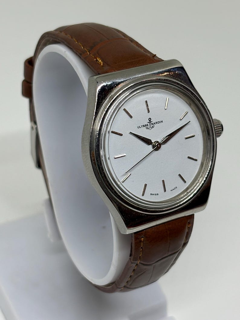 May include: A silver-tone wristwatch with a white dial featuring the Ulysse Nardin logo and anchor symbol. The watch has thin silver hour markers, silver hands, and a brown leather strap with a textured, embossed pattern and visible stitching.