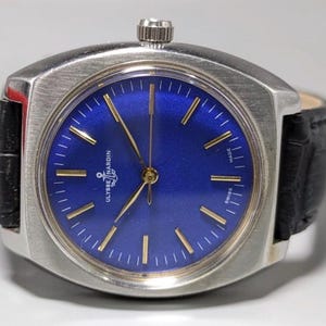 May include: A vintage Ulysse Nardin wristwatch with a square stainless steel case and a deep blue dial. The watch features gold-tone hands and markers, a black leather band, and the brand name is visible on the dial. The watch is marked "Swiss Made".