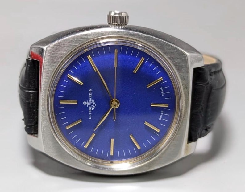 May include: A vintage Ulysse Nardin wristwatch with a square stainless steel case and a deep blue dial. The watch features gold-tone hands and markers, a black leather band, and the brand name is visible on the dial. The watch is marked "Swiss Made".