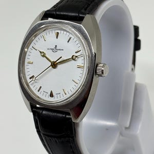 K&ouml;nnte beinhalten: Eine silberfarbene Armbanduhr mit wei&szlig;em Zifferblatt, goldfarbenen Zeigern und einem schwarzen Lederarmband mit Strukturmuster. Das Zifferblatt zeigt das Ulysse Nardin Logo mit Ankersymbol, goldene Stundenmarkierungen und den Schriftzug Swiss Made.