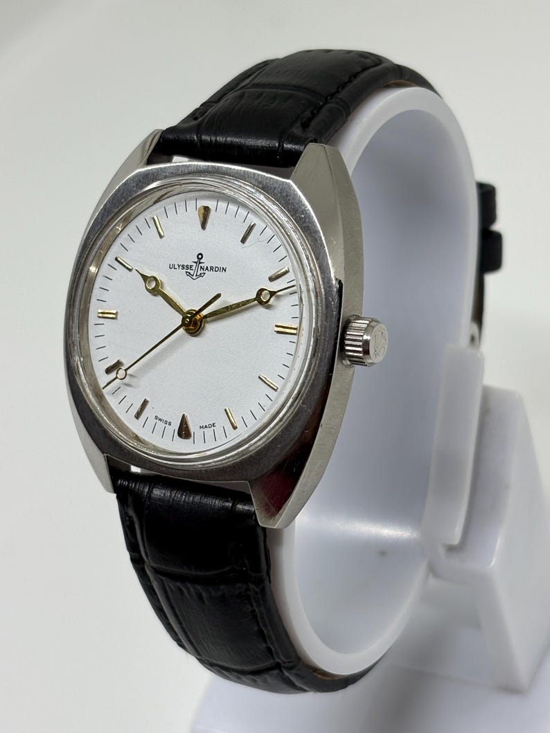 K&ouml;nnte beinhalten: Eine silberfarbene Armbanduhr mit wei&szlig;em Zifferblatt, goldfarbenen Zeigern und einem schwarzen Lederarmband mit Strukturmuster. Das Zifferblatt zeigt das Ulysse Nardin Logo mit Ankersymbol, goldene Stundenmarkierungen und den Schriftzug Swiss Made.