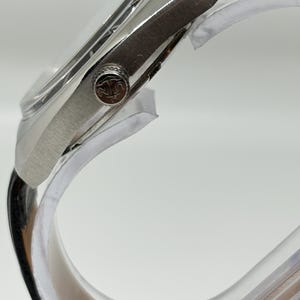 May include: A silver-toned wristwatch with a polished metal case and a black leather strap. The side of the watch features a circular crown embossed with a logo depicting an anchor and the letters U and N. The watch is mounted on a clear stand.