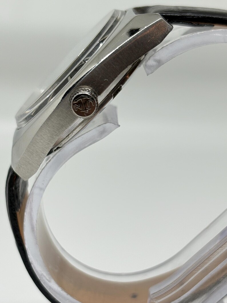 May include: A silver-toned wristwatch with a polished metal case and a black leather strap. The side of the watch features a circular crown embossed with a logo depicting an anchor and the letters U and N. The watch is mounted on a clear stand.