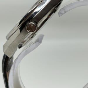 May include: A silver-toned wristwatch with a black leather strap. The side view shows a metallic case and a circular crown featuring the embossed letters JN. The watch is positioned on a clear curved display stand, highlighting the side profile.