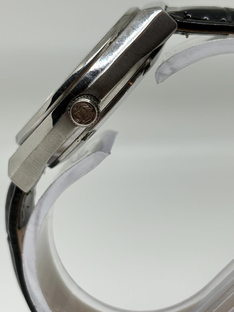 May include: A silver-toned wristwatch with a black leather strap. The side view shows a metallic case and a circular crown featuring the embossed letters JN. The watch is positioned on a clear curved display stand, highlighting the side profile.
