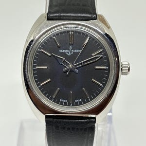 May include: A silver-tone wristwatch with a round black dial, silver hour markers, and silver hands. The watch features the Ulysse Nardin logo with an anchor symbol at the top and the text SWISS MADE at the bottom. It has a black leather strap.