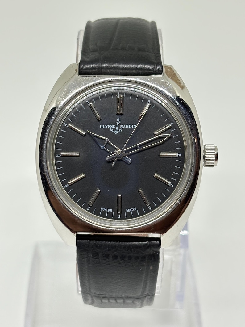 May include: A silver-tone wristwatch with a round black dial, silver hour markers, and silver hands. The watch features the Ulysse Nardin logo with an anchor symbol at the top and the text SWISS MADE at the bottom. It has a black leather strap.