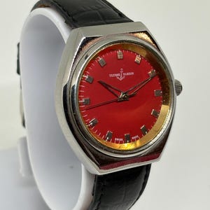 May include: A Ulysse Nardin wristwatch featuring a vibrant red dial with silver-tone hour markers and hands. The watch has a polished silver-tone metal case and a black leather strap with a textured finish. The dial displays the brand name and logo.