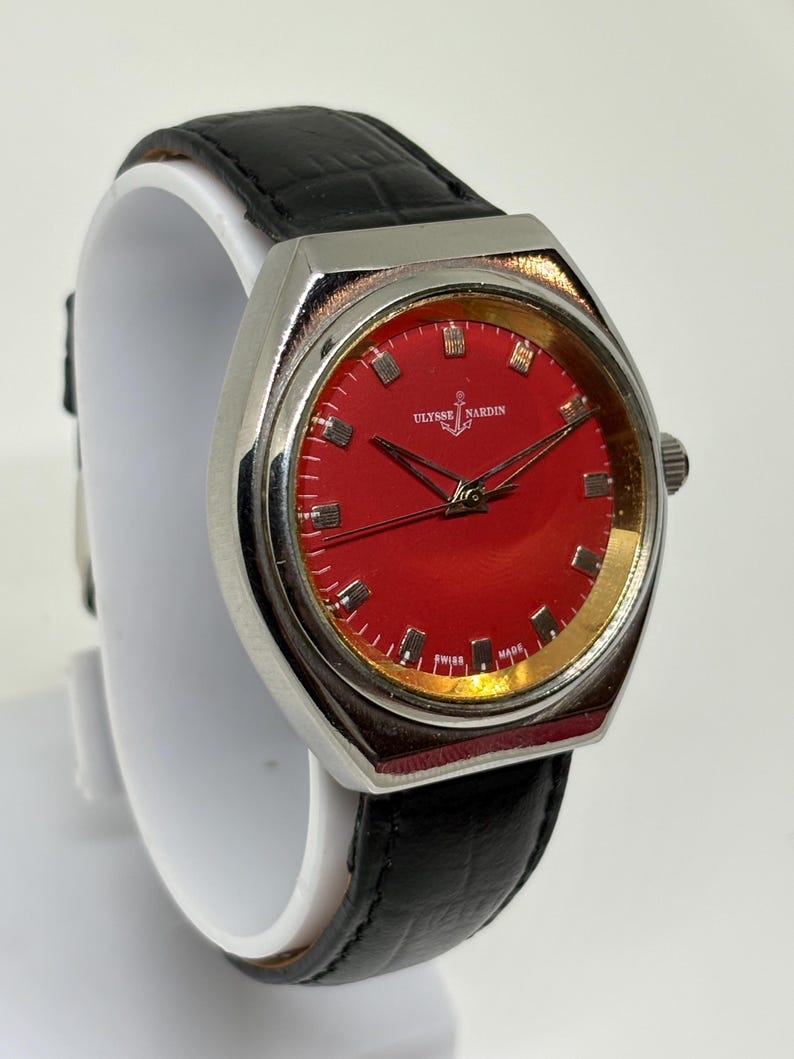 May include: A Ulysse Nardin wristwatch featuring a vibrant red dial with silver-tone hour markers and hands. The watch has a polished silver-tone metal case and a black leather strap with a textured finish. The dial displays the brand name and logo.