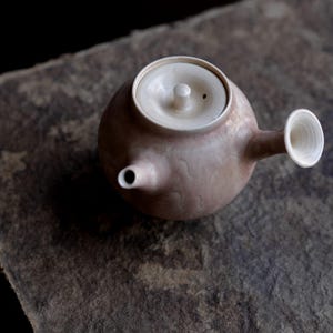 May include: A small, handcrafted ceramic teapot with a light brown body and a white lid and spout. The teapot has a rounded shape and a small handle. The background is a textured, gray surface.